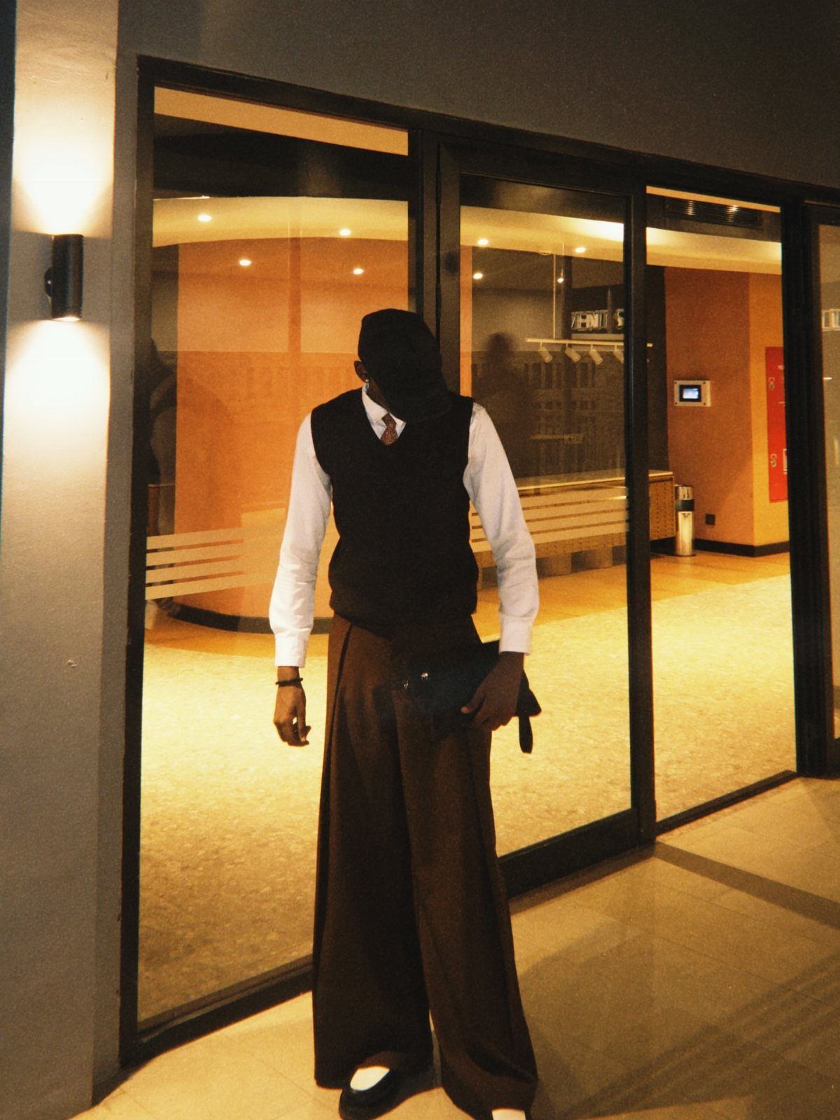 Person wearing a brown vest, white shirt, and wide-leg brown trousers standing outdoors.