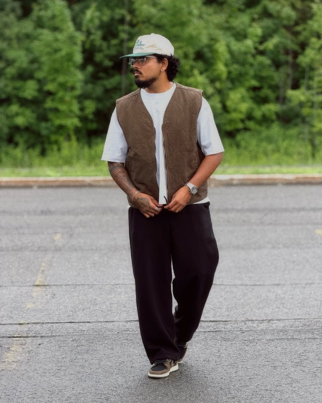 Man wearing a brown vest, white t-shirt, and black trousers in a parking lot