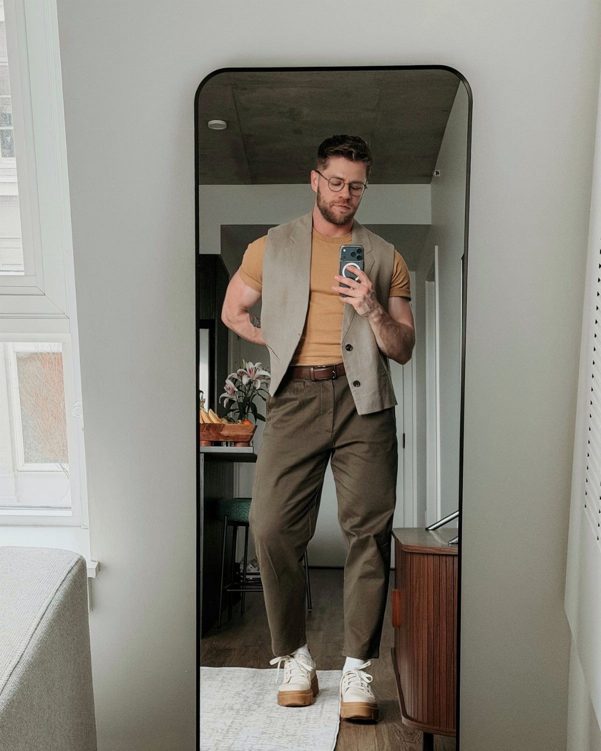 Man in a tan vest and olive brown trousers taking a mirror selfie.