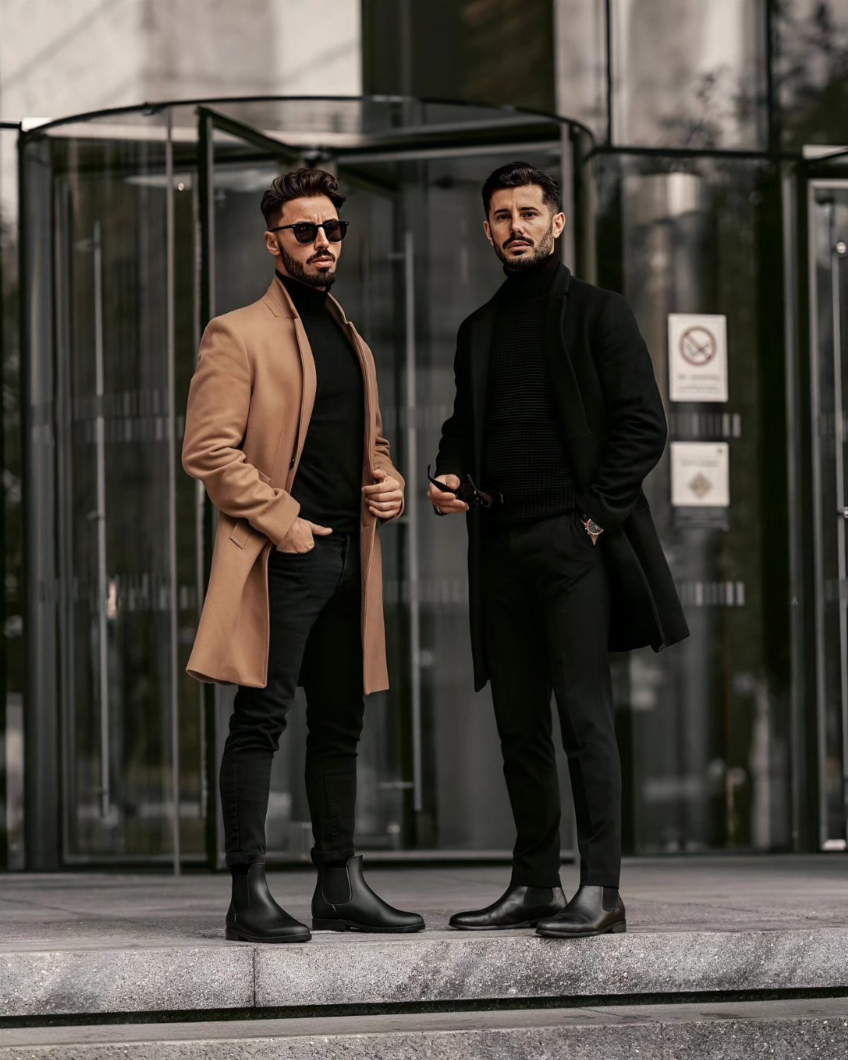 Two men in coats and black outfits posing in front of glass doors.