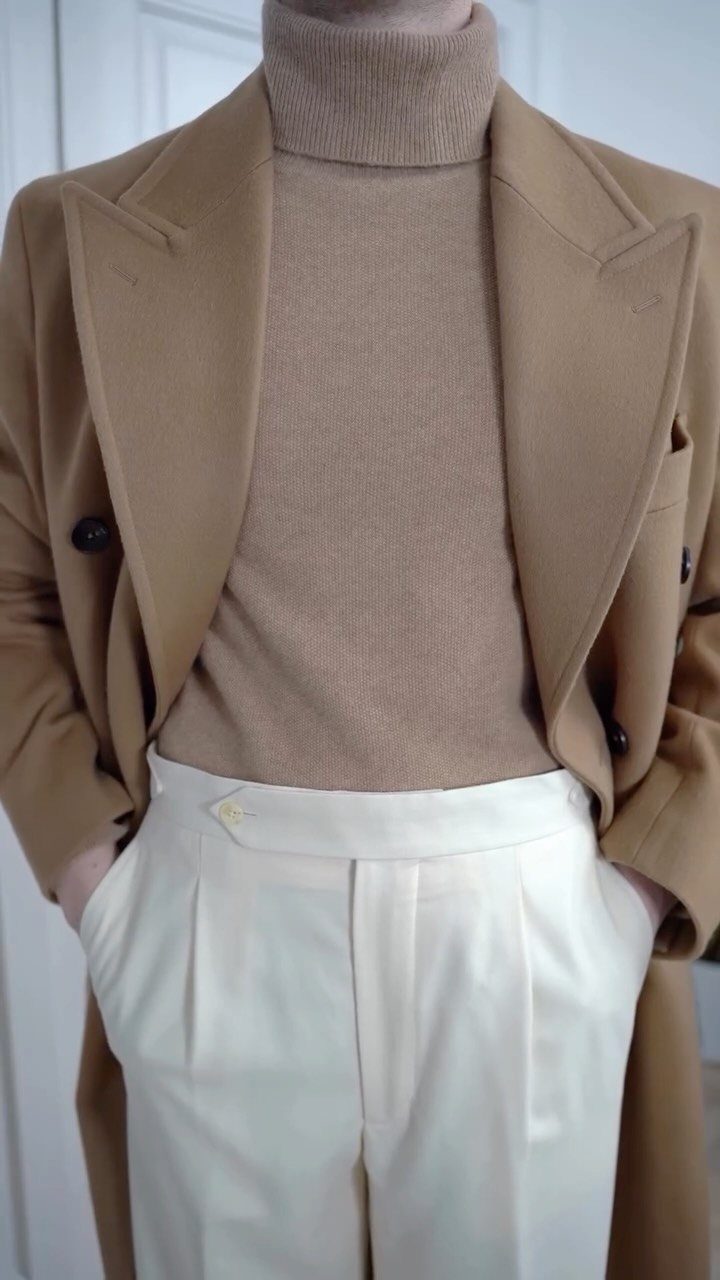 Person wearing a camel coat, beige turtleneck, and off-white pleated trousers indoors.