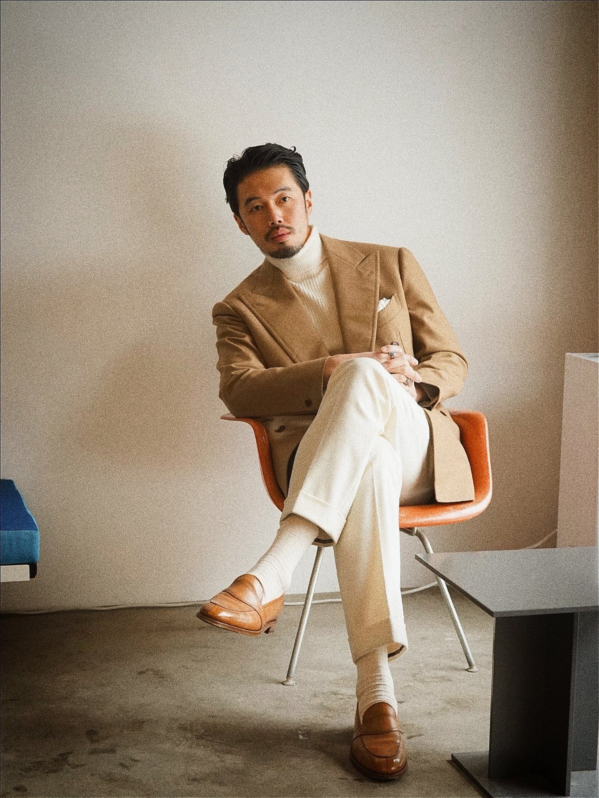 Man wearing a camel coat with a cream turtleneck and off-white trousers sitting.