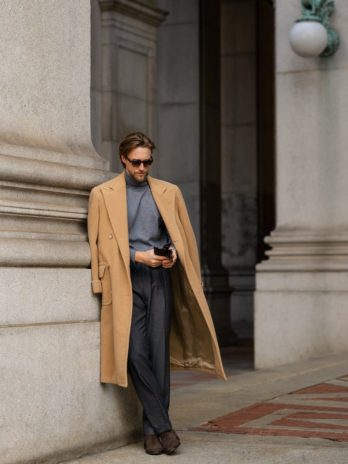 Man in a camel coat, light blue turtleneck, and grey trousers leaning on stone.