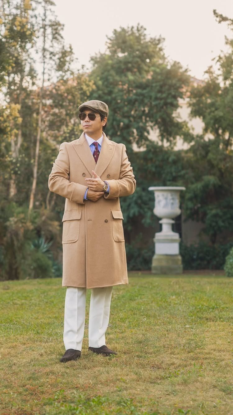 Man in camel overcoat and white trousers posing in a green garden setting