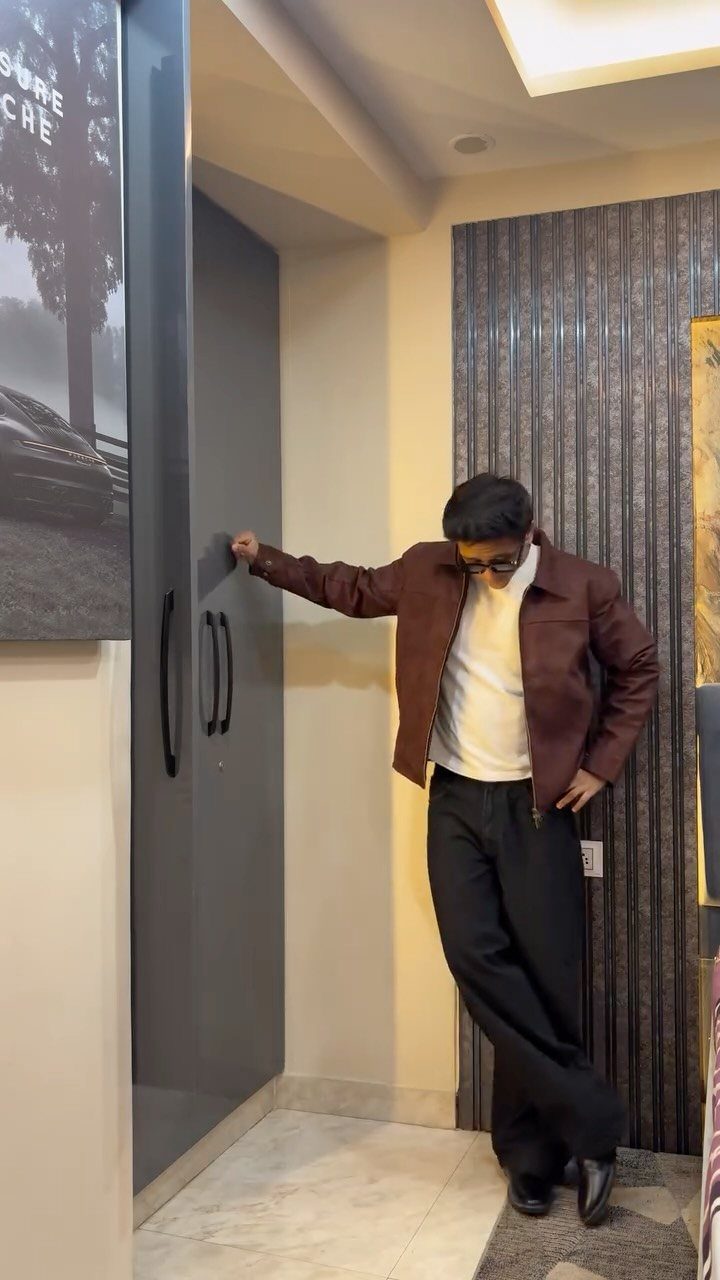Man wearing a brown trucker jacket, white t-shirt, and black trousers indoors.
