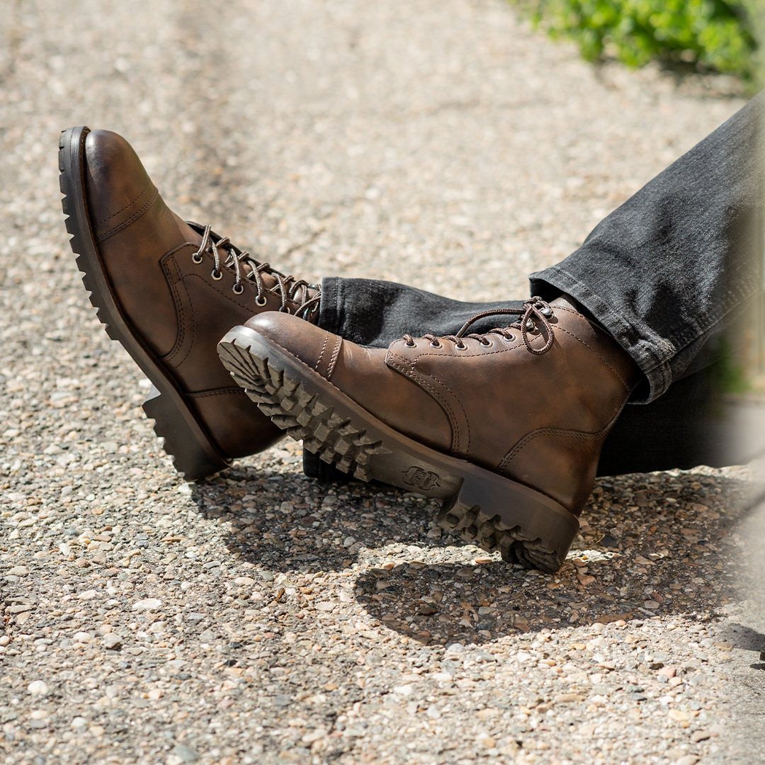 Brown leather lace-up work boots worn with black jeans on gravel ground.