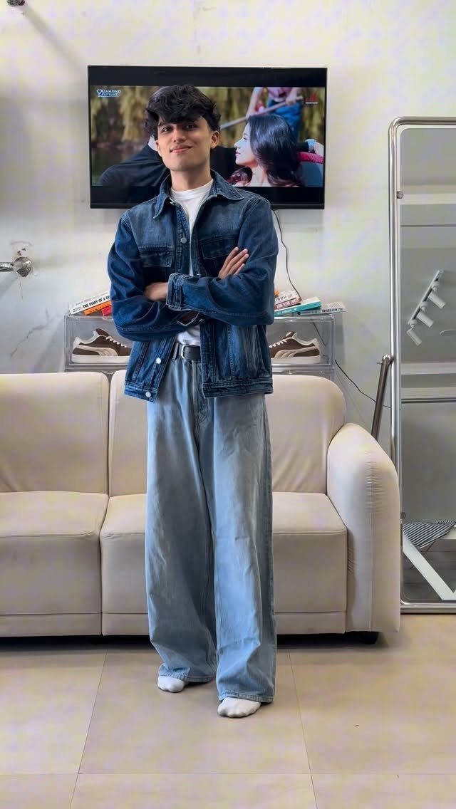 Young man wearing a denim jacket and wide-leg light blue denim jeans indoors.