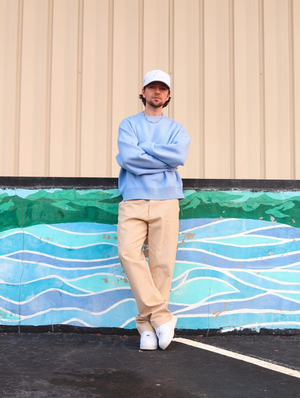 Man wearing a pastel blue sweater and tan chinos standing by a mural.