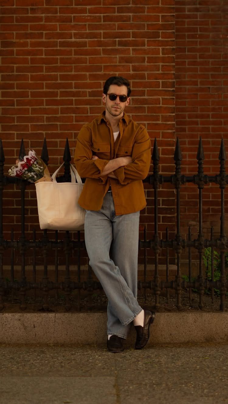 Man in mustard shirt and light blue jeans leaning against a brick wall