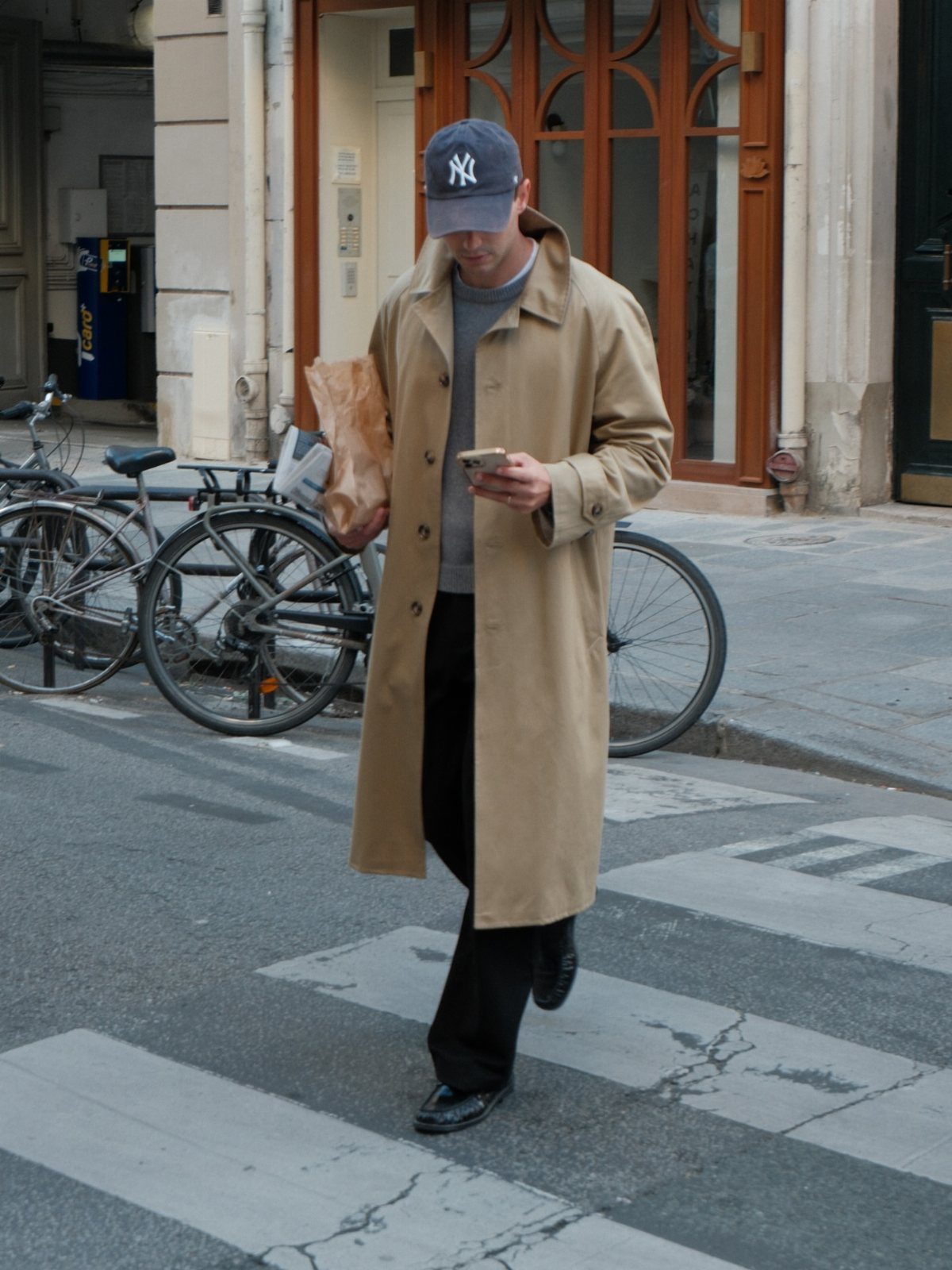 Man in a tan trench coat and blue cap walking across a city street