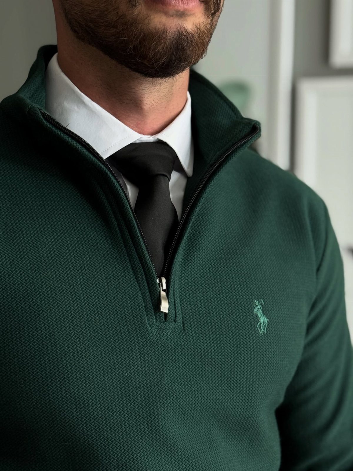 Man wearing a dark green quarter-zip sweater over a white shirt and black tie.