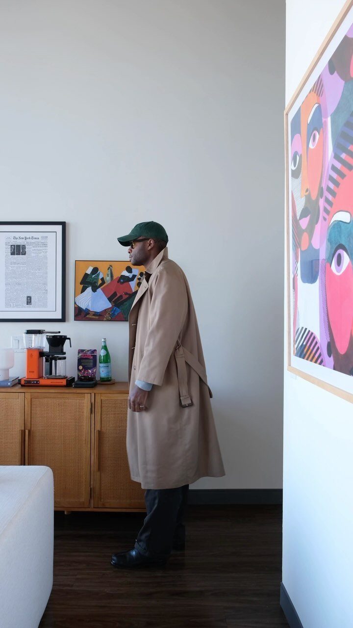 Man in a beige trench coat and green cap standing in a modern room.