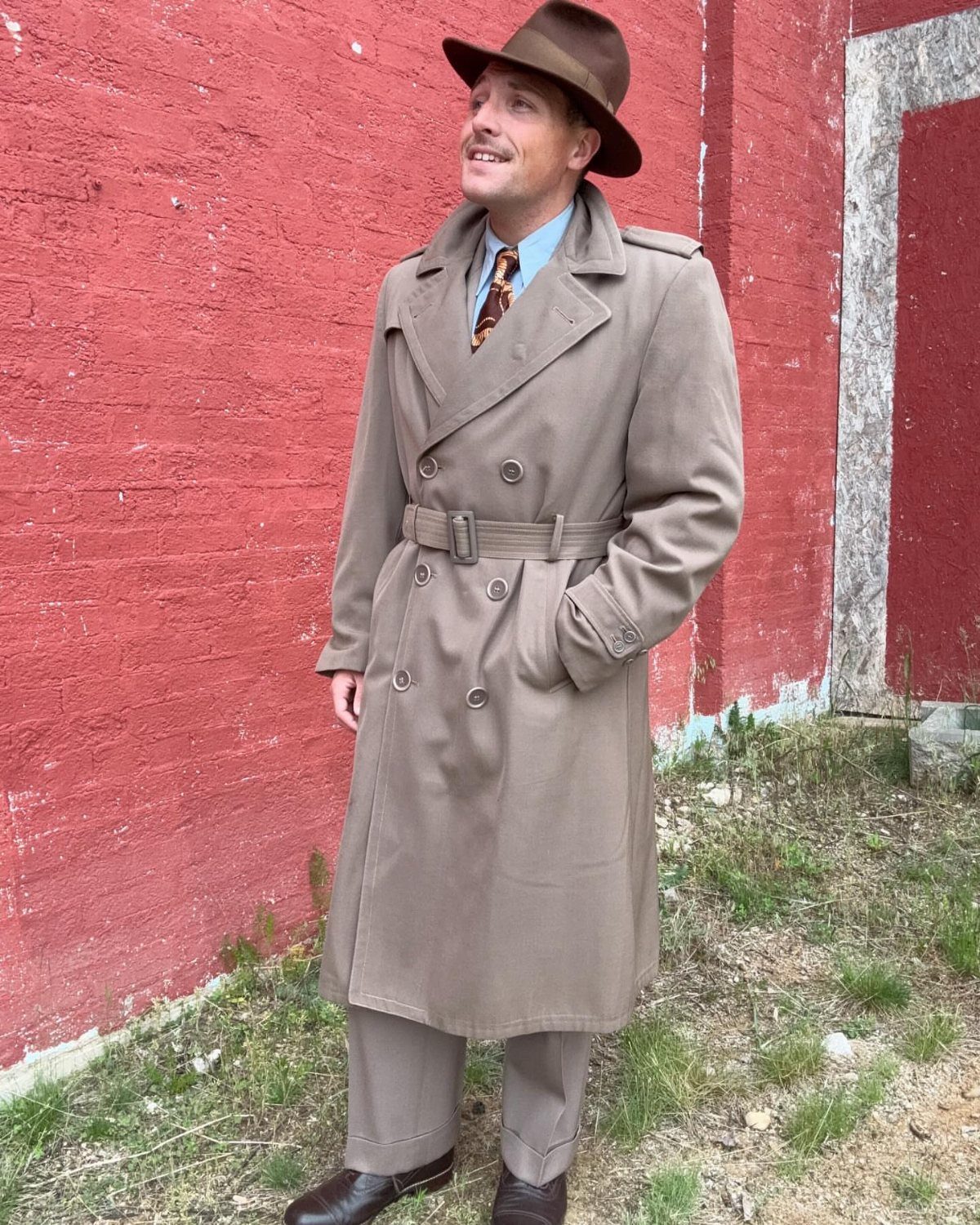 Man in a khaki trench coat and brown fedora posing by red wall