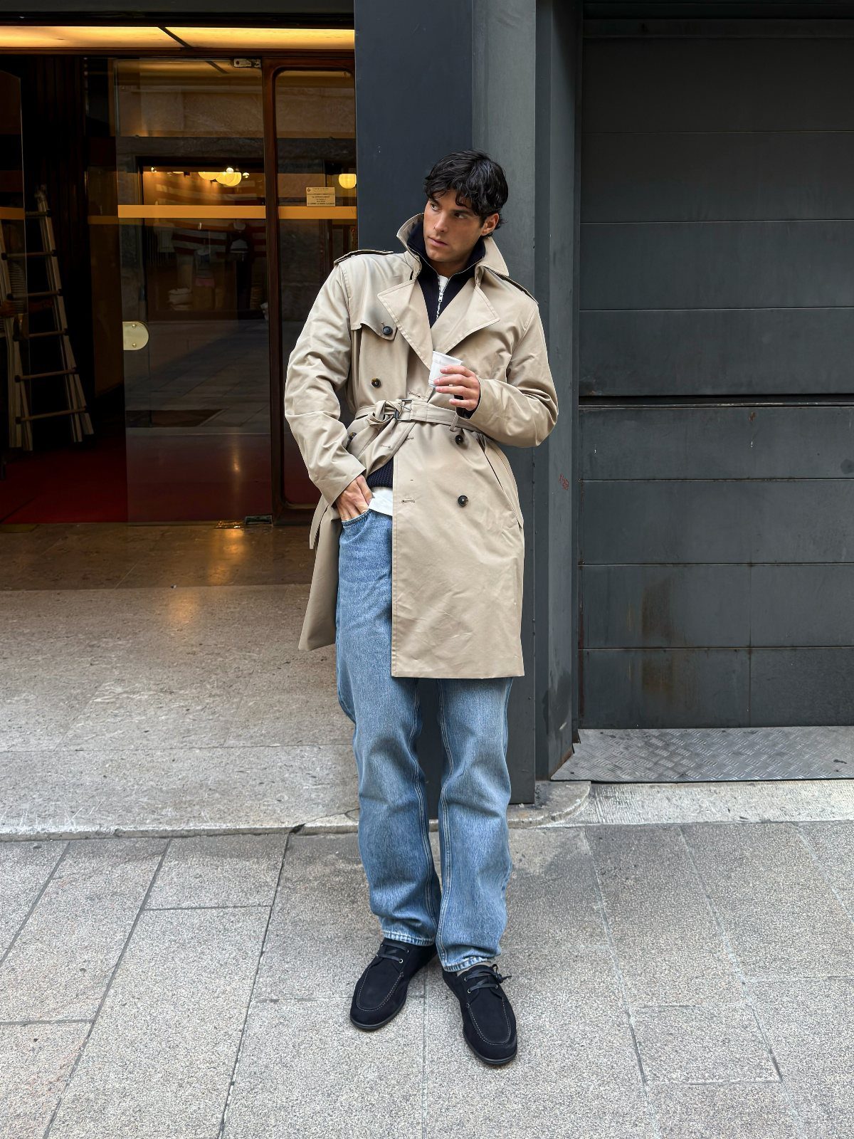 Man wearing a beige trench coat and light blue jeans standing by a wall