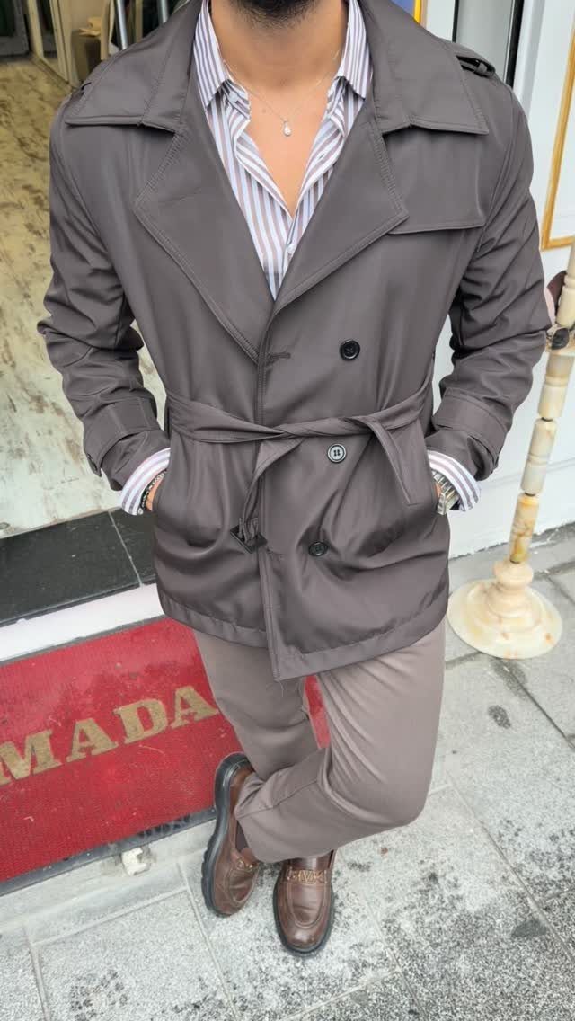 Man wearing a brown belted trench coat, striped shirt, and brown trousers.