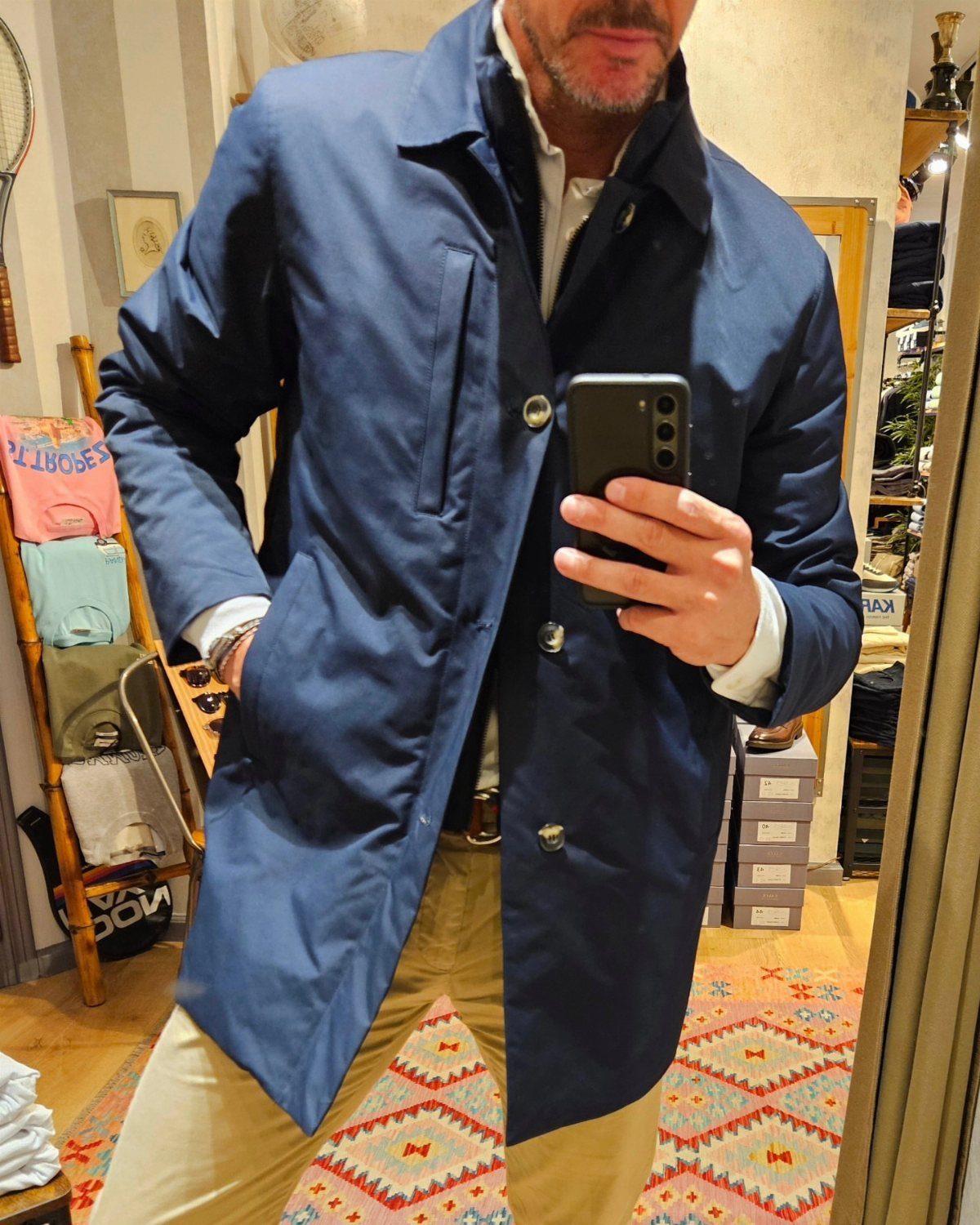 Man in navy blue trench coat and khaki pants taking a mirror selfie.