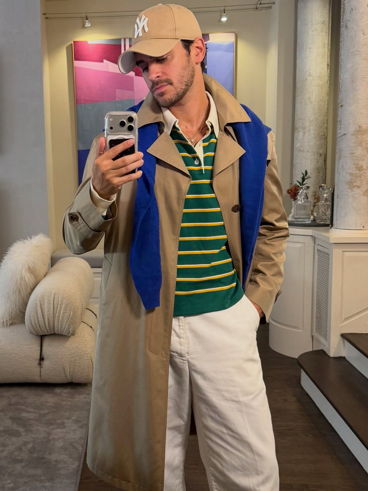 Man in tan trench coat, striped polo, and baseball cap taking a mirror selfie.