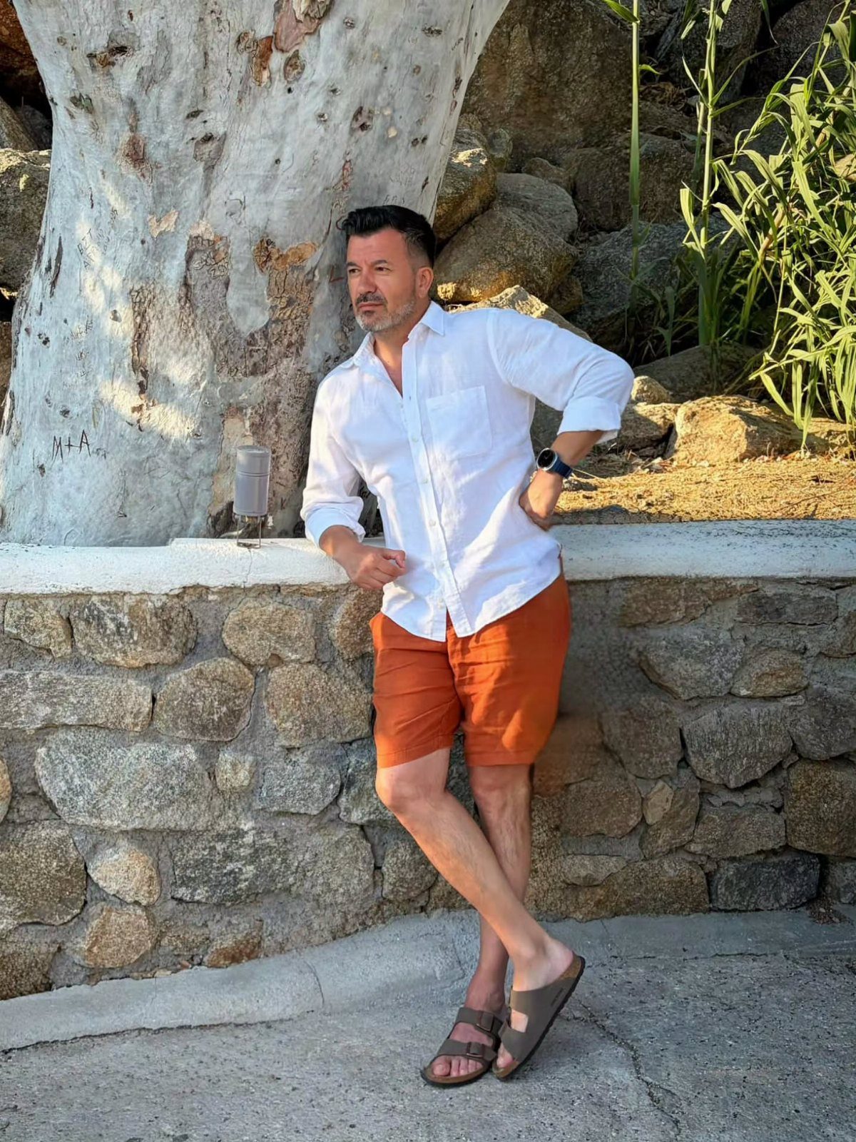 Man wearing a white button-down shirt and orange tailored shorts standing by a wall
