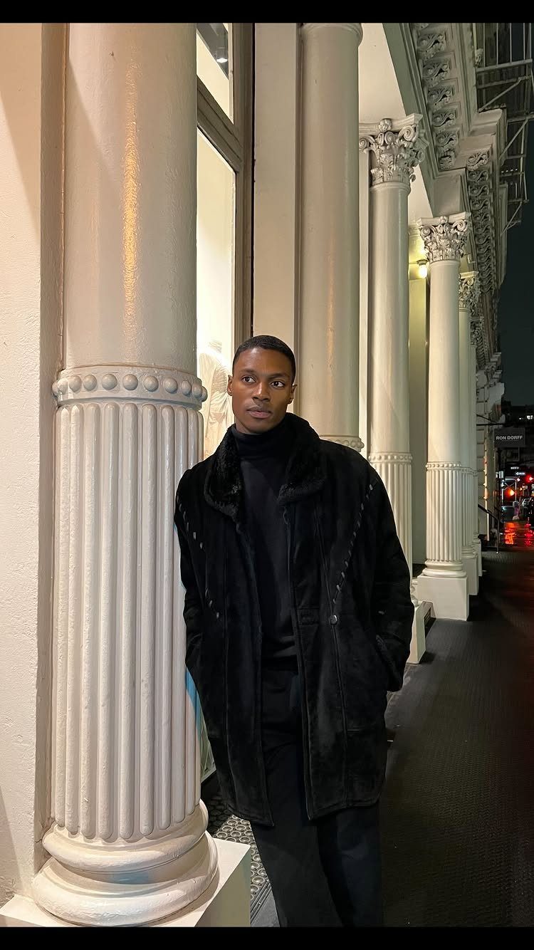 Man in a black winter coat standing by white classical columns at night.