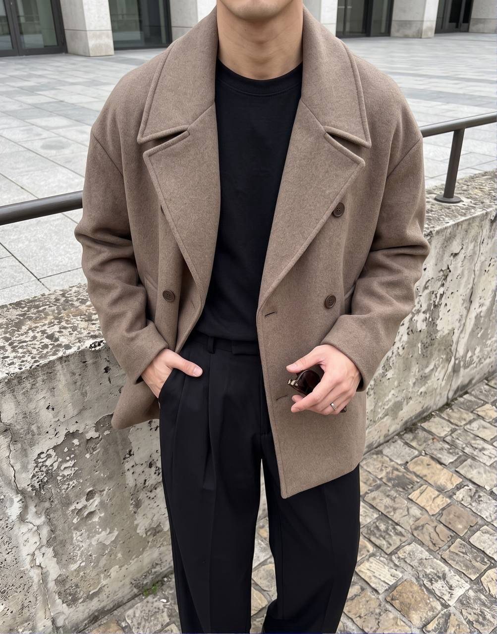 Man wearing a tan double-breasted peacoat with black trousers on a city street.