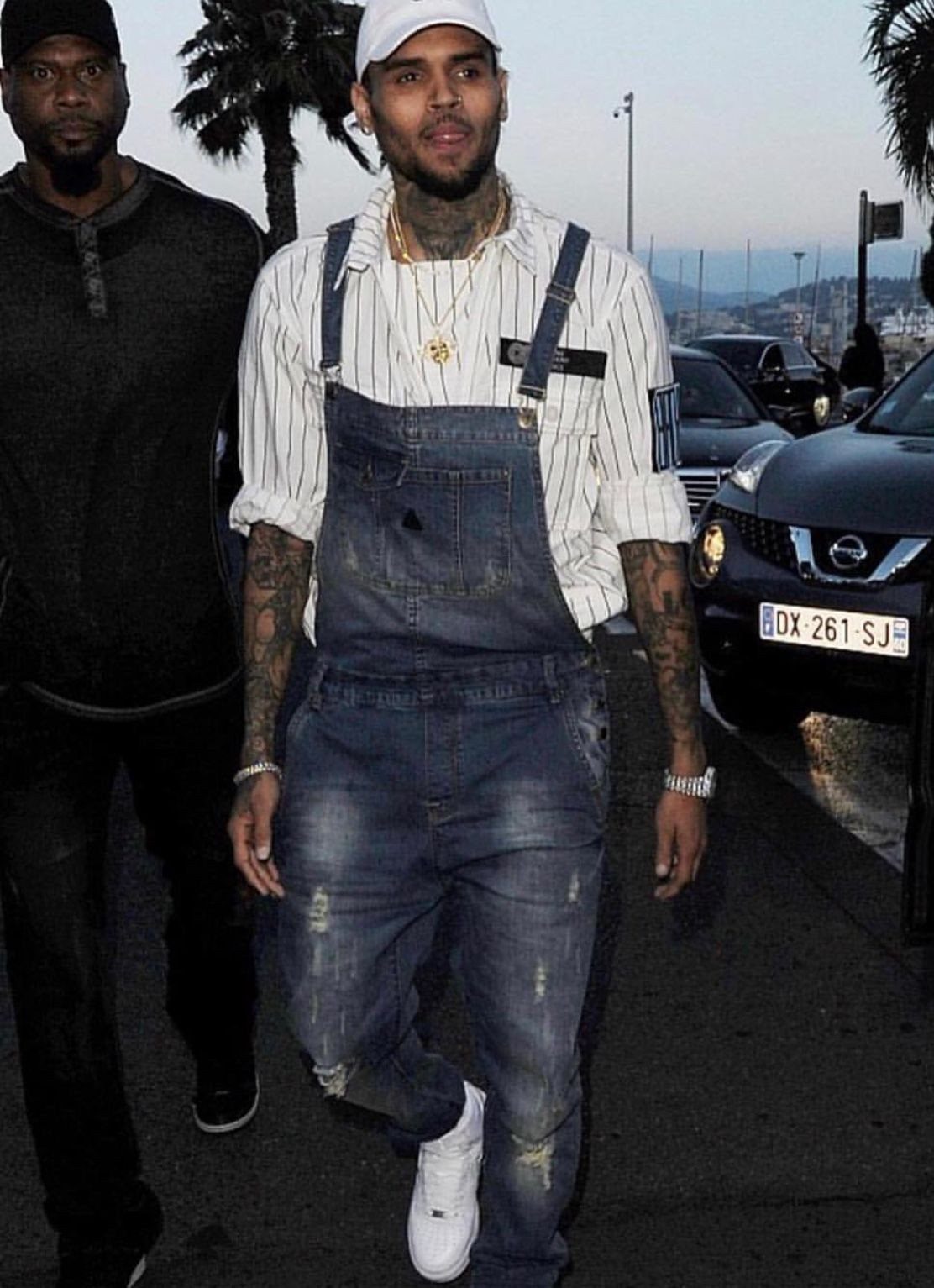Chris Brown wearing baggy blue denim overalls and a white pinstripe shirt outdoors.