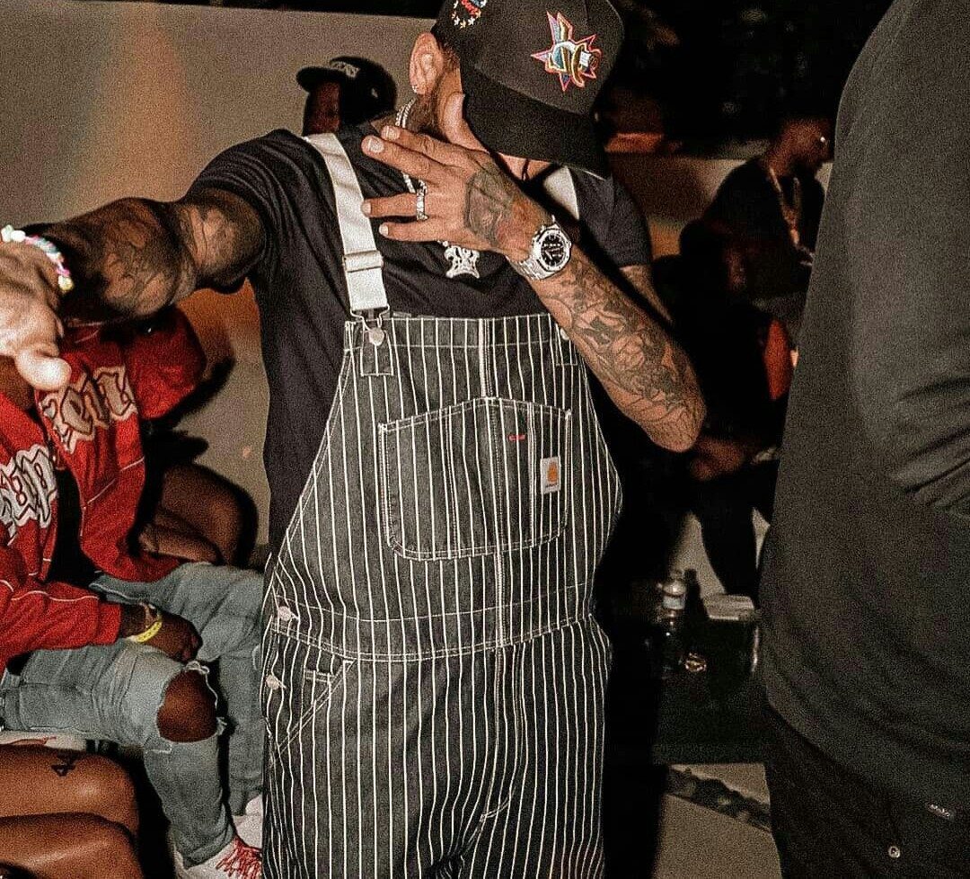 Man wearing black pinstripe overalls and a black t-shirt with a baseball cap.