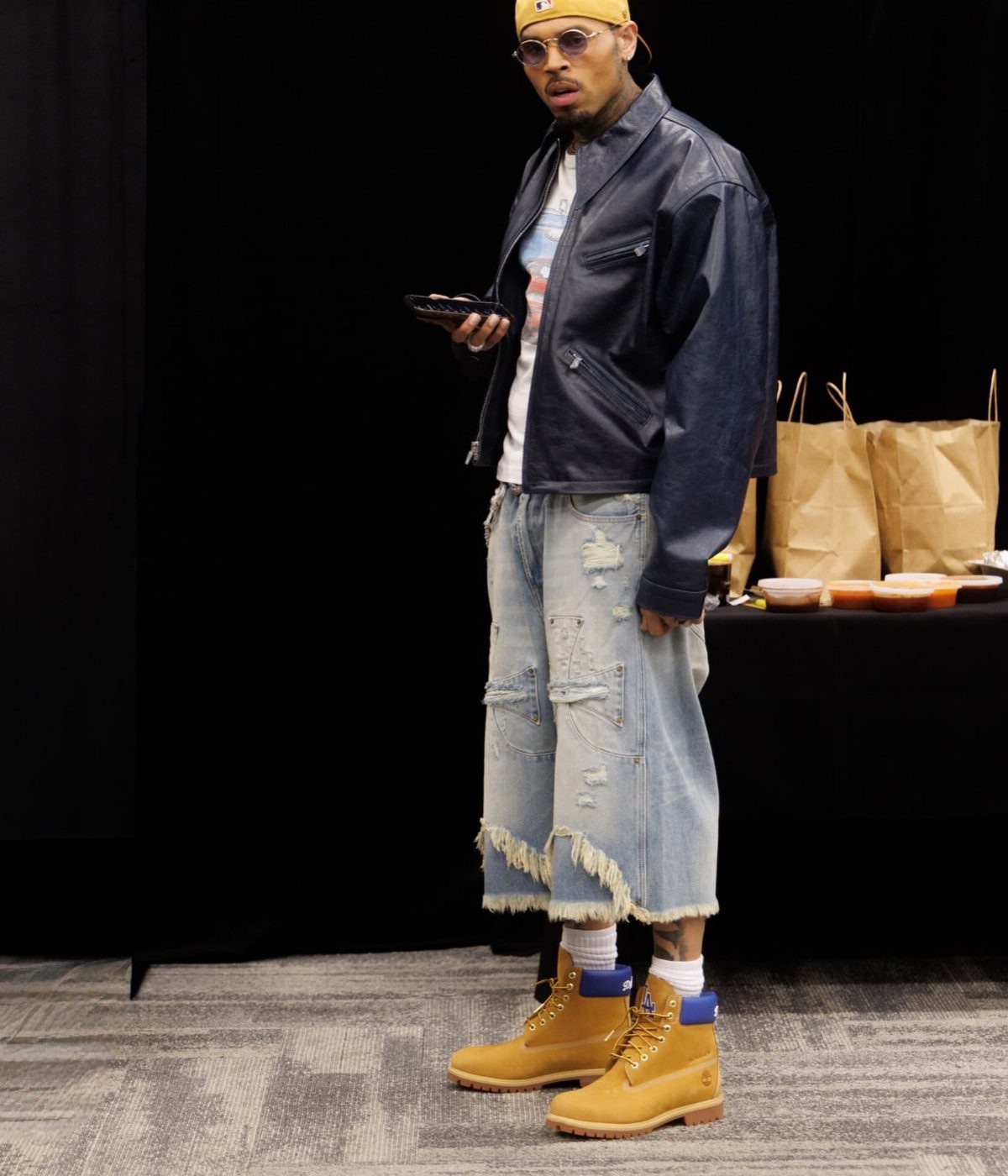 Man wearing light blue distressed denim overalls, black leather jacket, and tan boots.