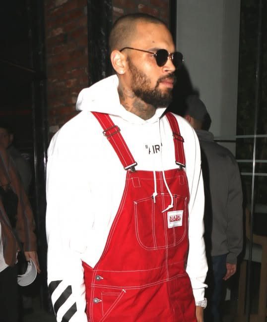 Man wearing red denim overalls over a white hooded sweatshirt and dark sunglasses.