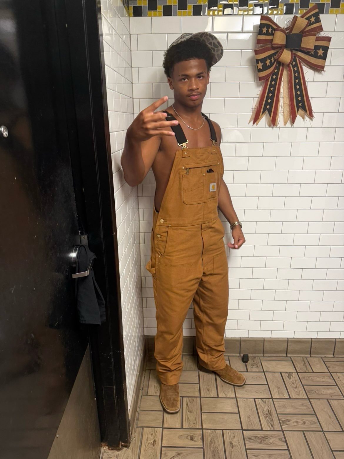 Man in brown canvas overalls and a patterned cap posing against white tiles.