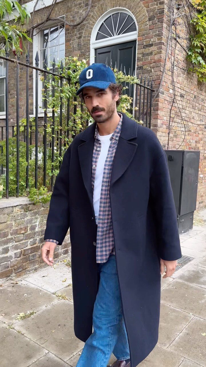 Man wearing a long navy overcoat and blue jeans standing on a city sidewalk.