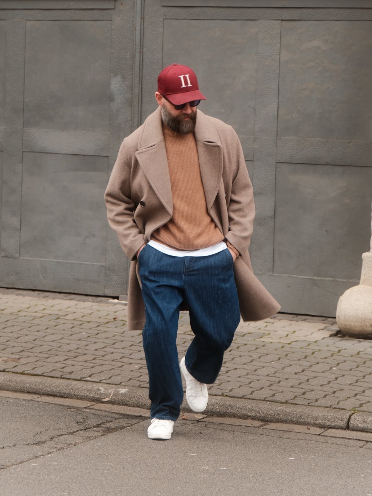 Man wearing a beige overcoat and blue jeans walking on a city street.