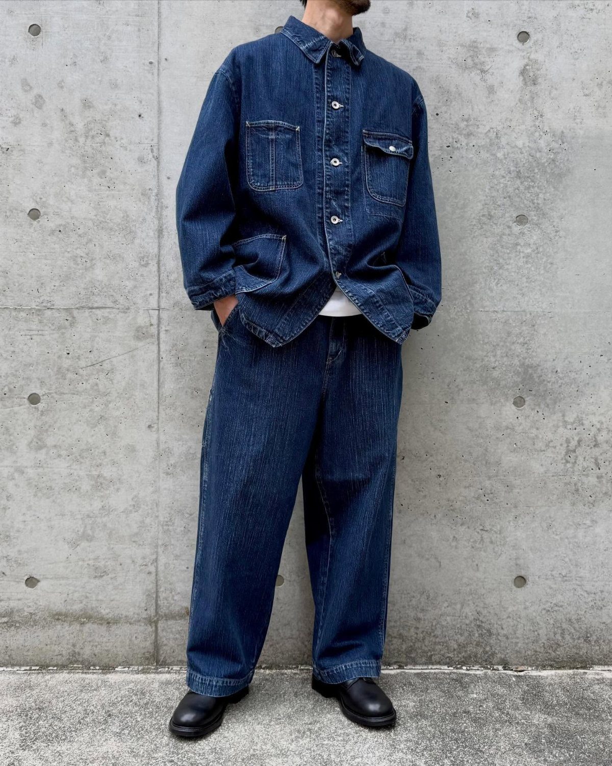 Person wearing a dark blue denim jacket and wide-leg indigo jeans against concrete.