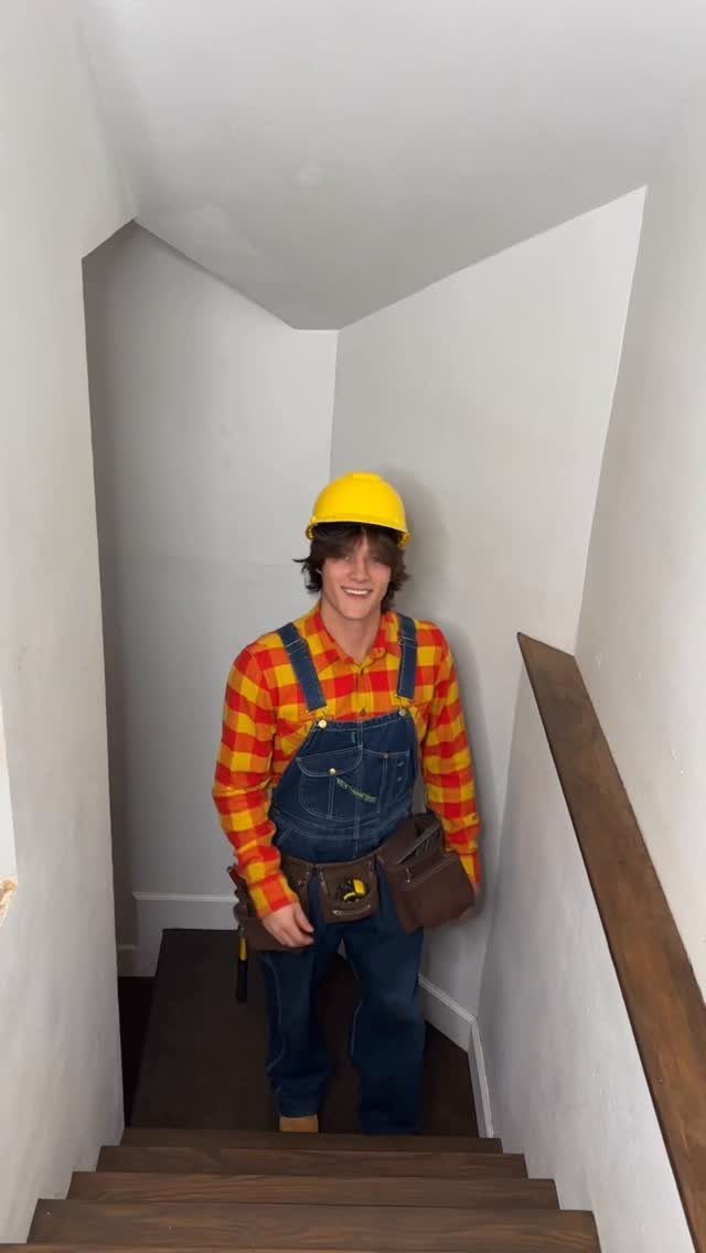 Man in construction worker costume wearing blue overalls, plaid shirt, and yellow hard hat.