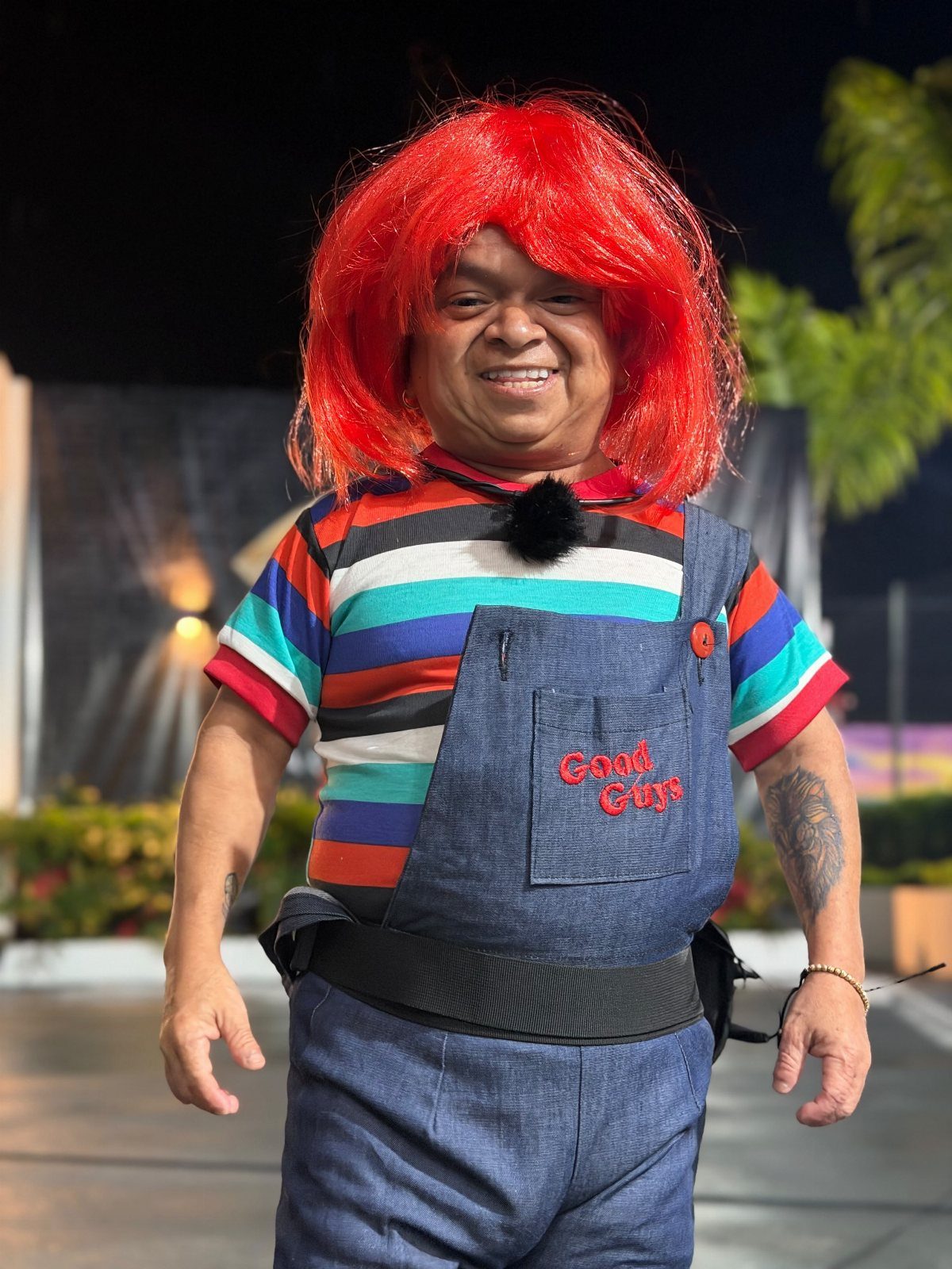 Person in Chucky costume with red wig, striped shirt, and Good Guys overalls.