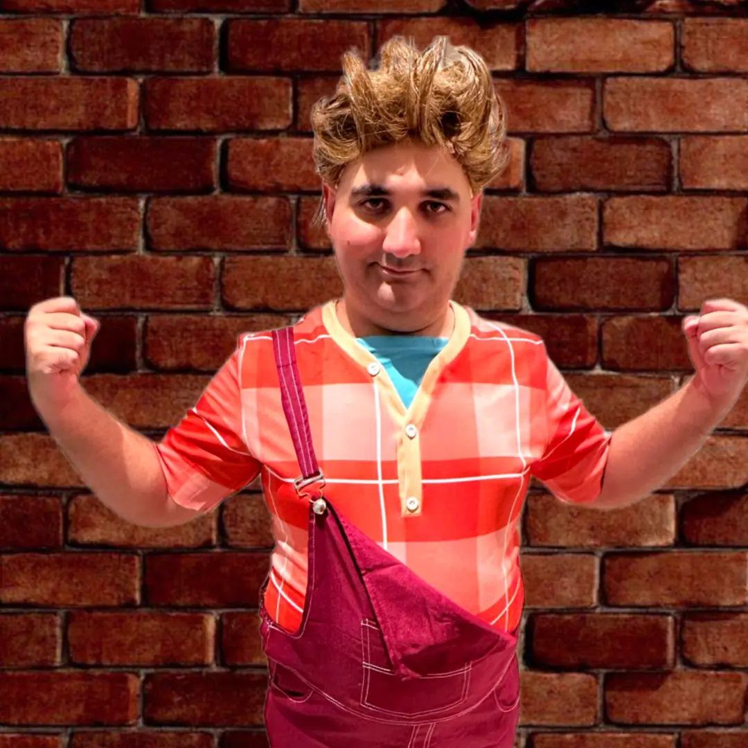 Man in red plaid shirt and maroon overalls posing against a brick wall.