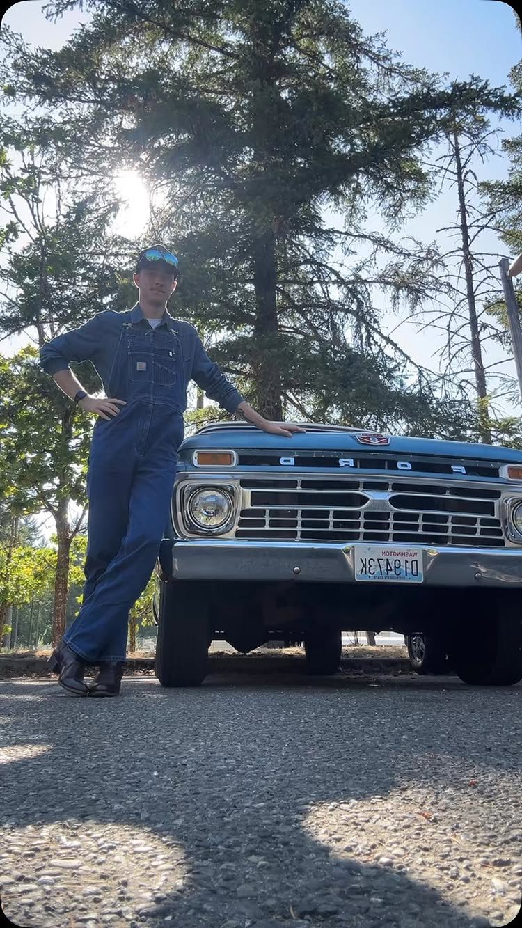 Man in blue denim overalls posing next to a vintage light-blue pickup truck.