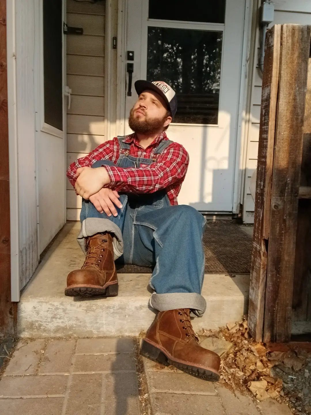 Man in red checkered shirt and blue overalls wearing brown leather boots.