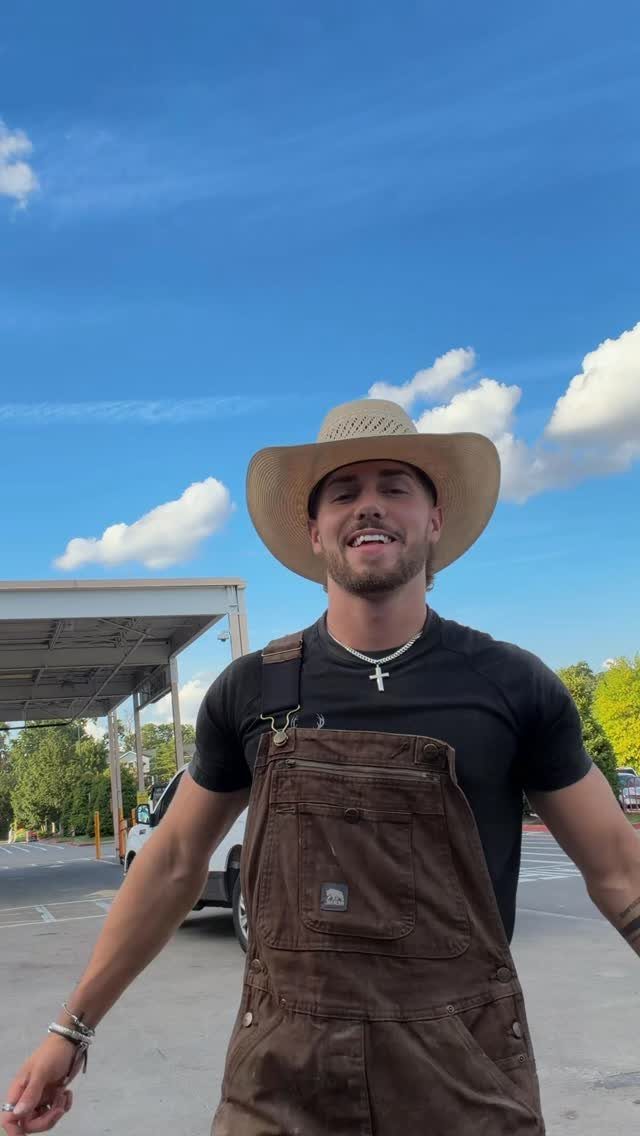 Man wearing brown canvas overalls, a black t-shirt, and a straw cowboy hat.