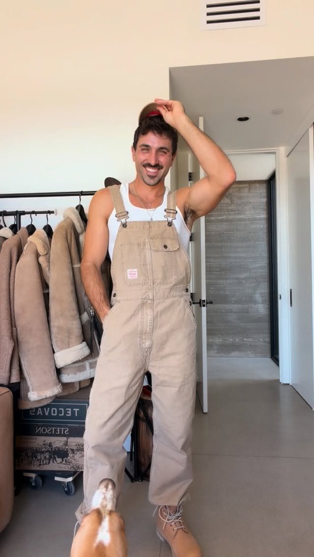 Man wearing tan overalls and a white tank top posing in a room
