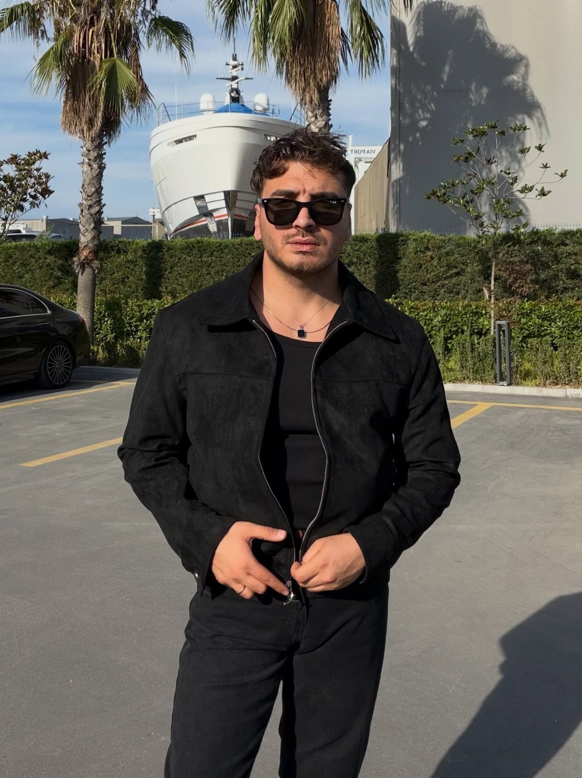 Man in a black suede jacket and black trousers standing in a parking lot