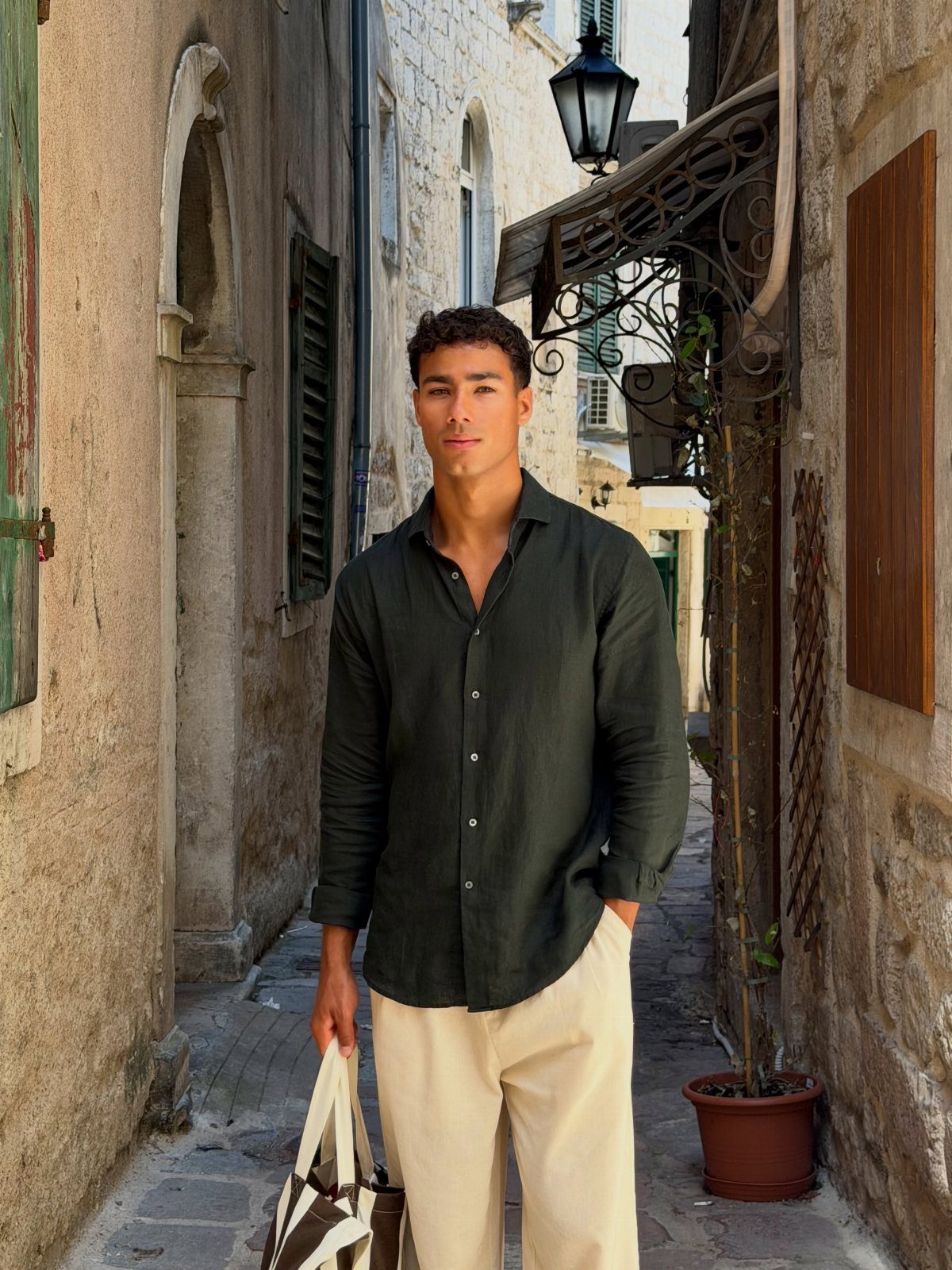 Man in a dark green long-sleeve shirt and cream trousers in a stone alley.