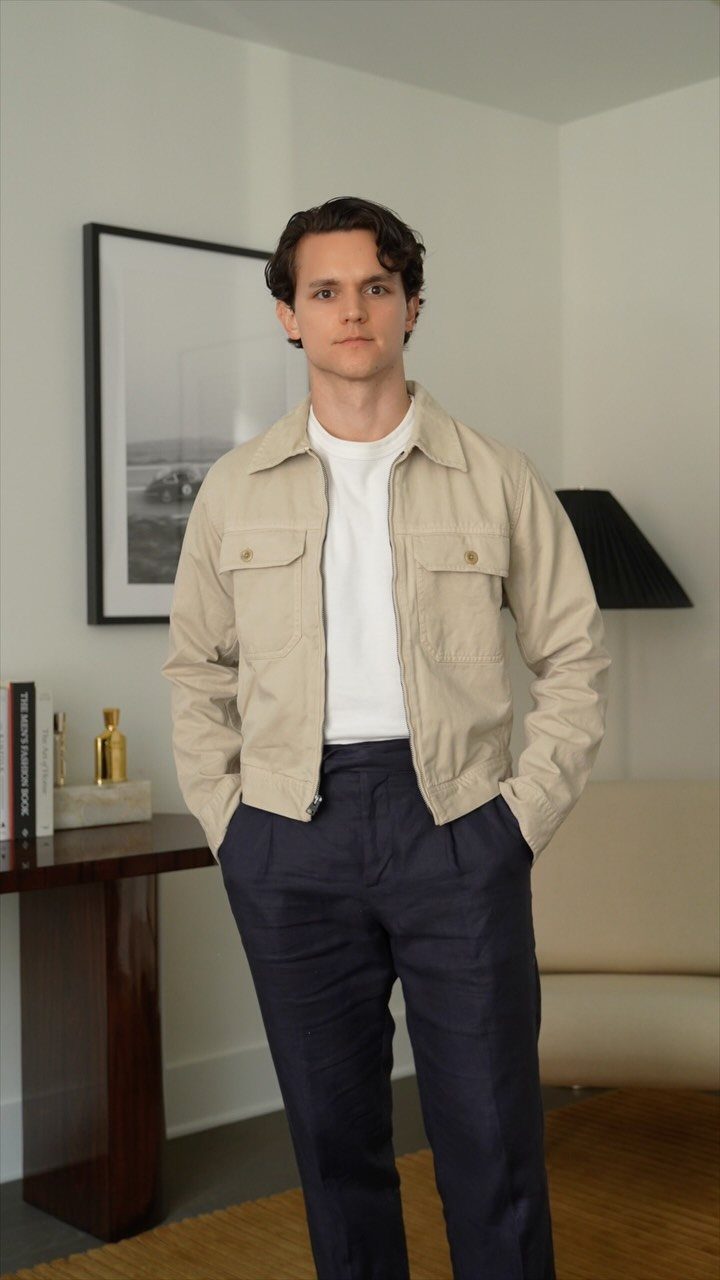 Man wearing a beige summer jacket, white t-shirt, and navy blue trousers indoors.