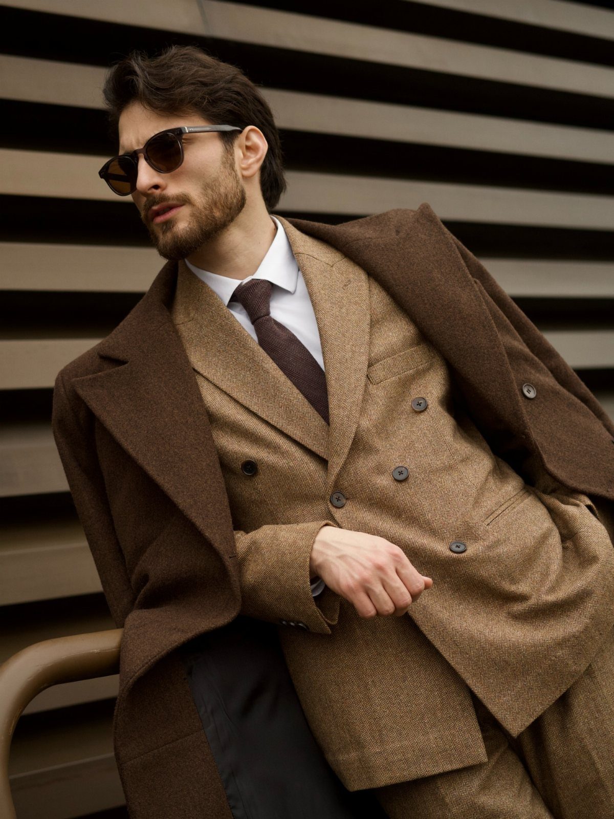 Man in a tan double-breasted suit and dark brown overcoat wearing sunglasses.