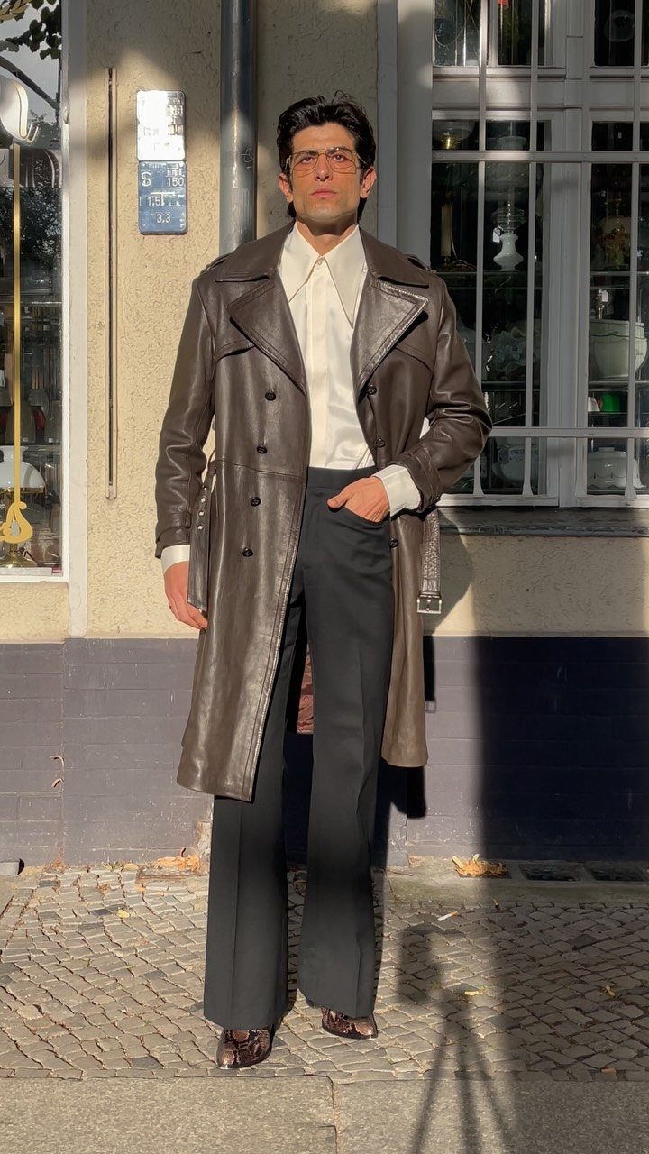 Man wearing a dark brown leather trench coat with black trousers and white shirt.
