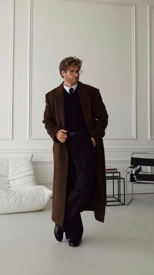 Man wearing a long dark brown overcoat with black trousers and a tie.