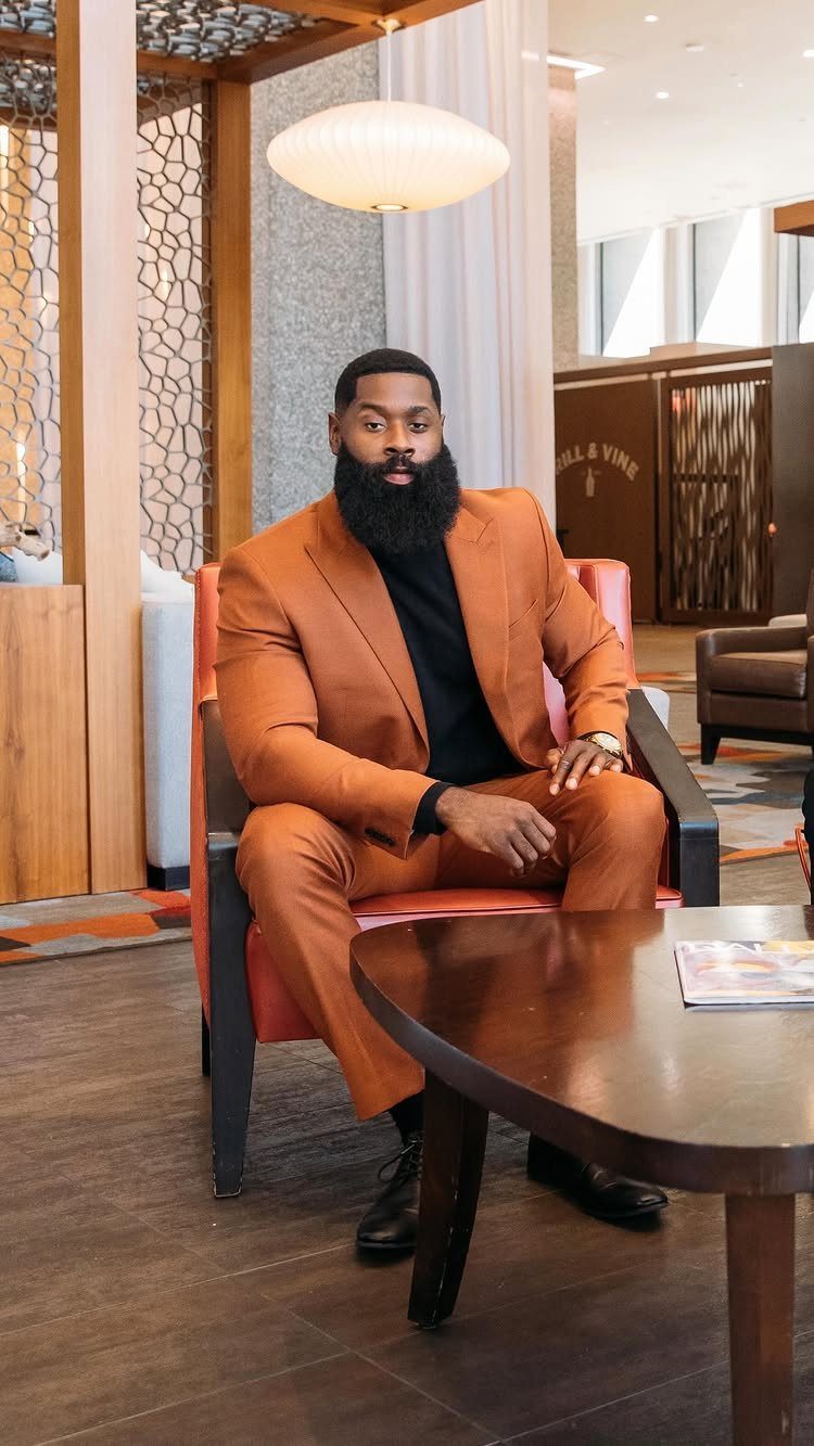 Man with a beard wearing a burnt orange suit and black shirt sitting indoors.