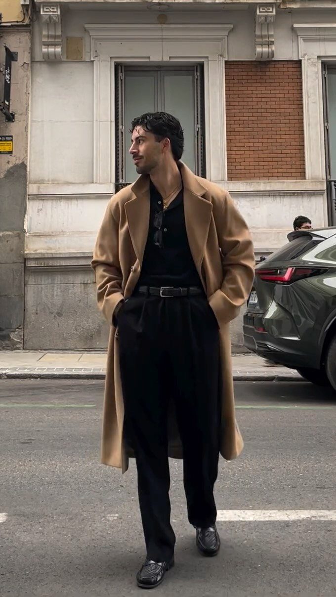 Man in a camel overcoat and black outfit standing on a city street.