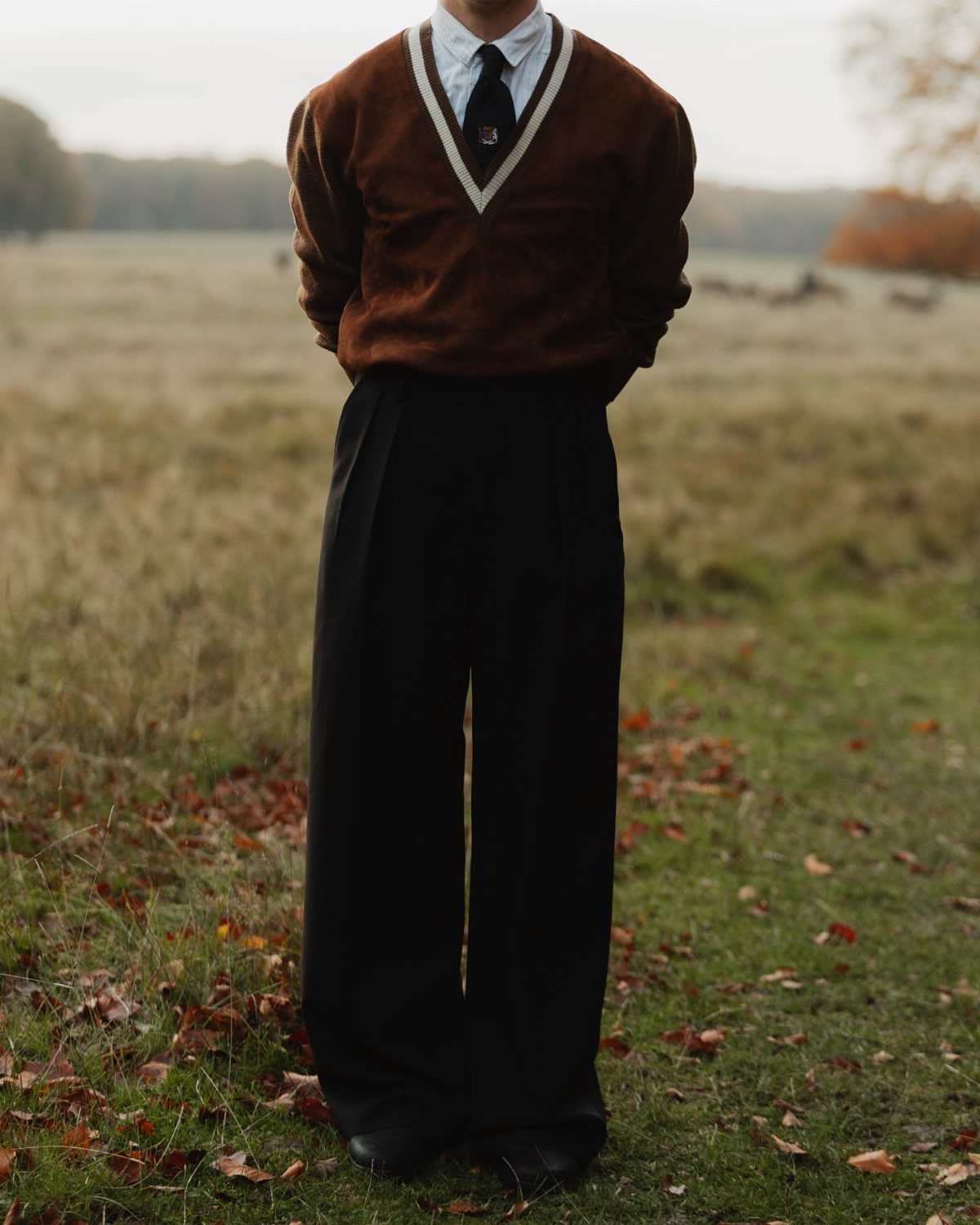 Person wearing a brown sweater, dark tie, and black trousers in an autumn field.