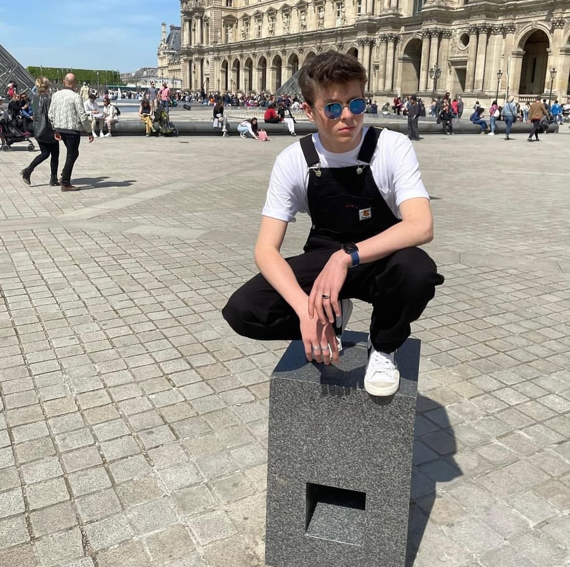 Man wearing black denim overalls and a white t-shirt sitting on stone plinth.