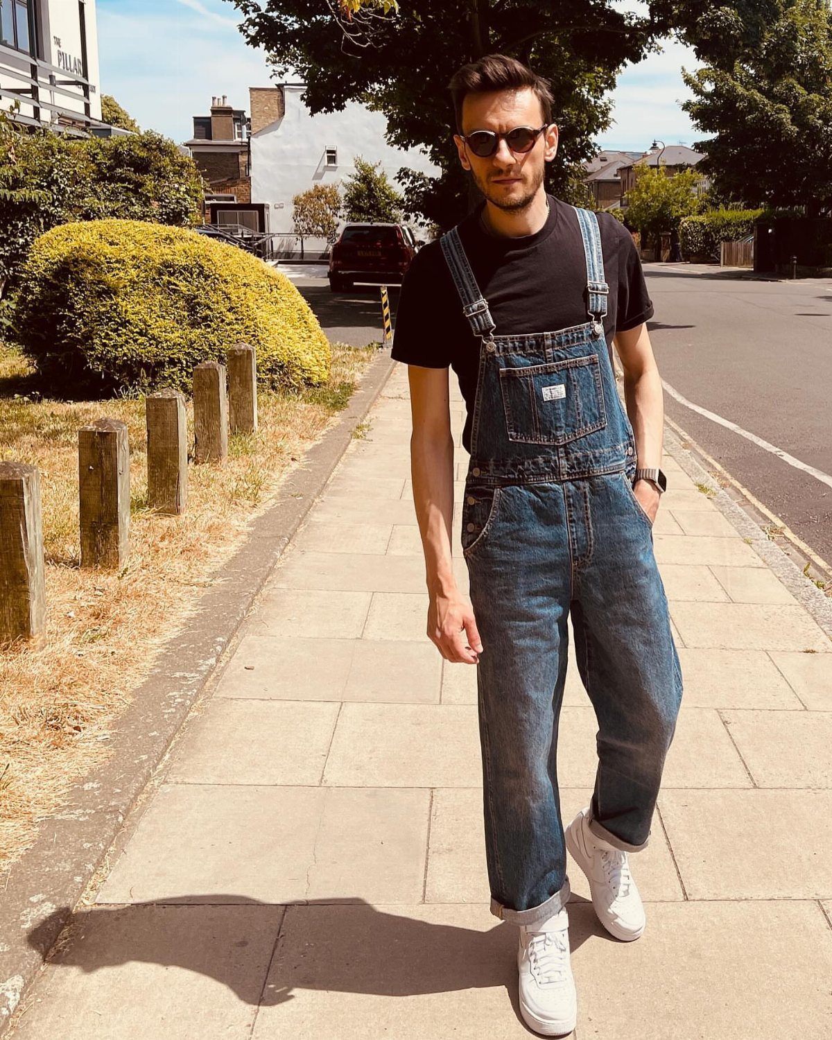 Man wearing blue denim overalls, a black t-shirt, and white sneakers on a sidewalk.